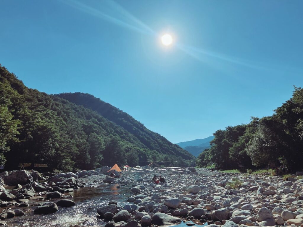 川辺の石と山、空に輝く太陽|自然の中でゆっくり呼吸を整える時間をイメージした山の風景